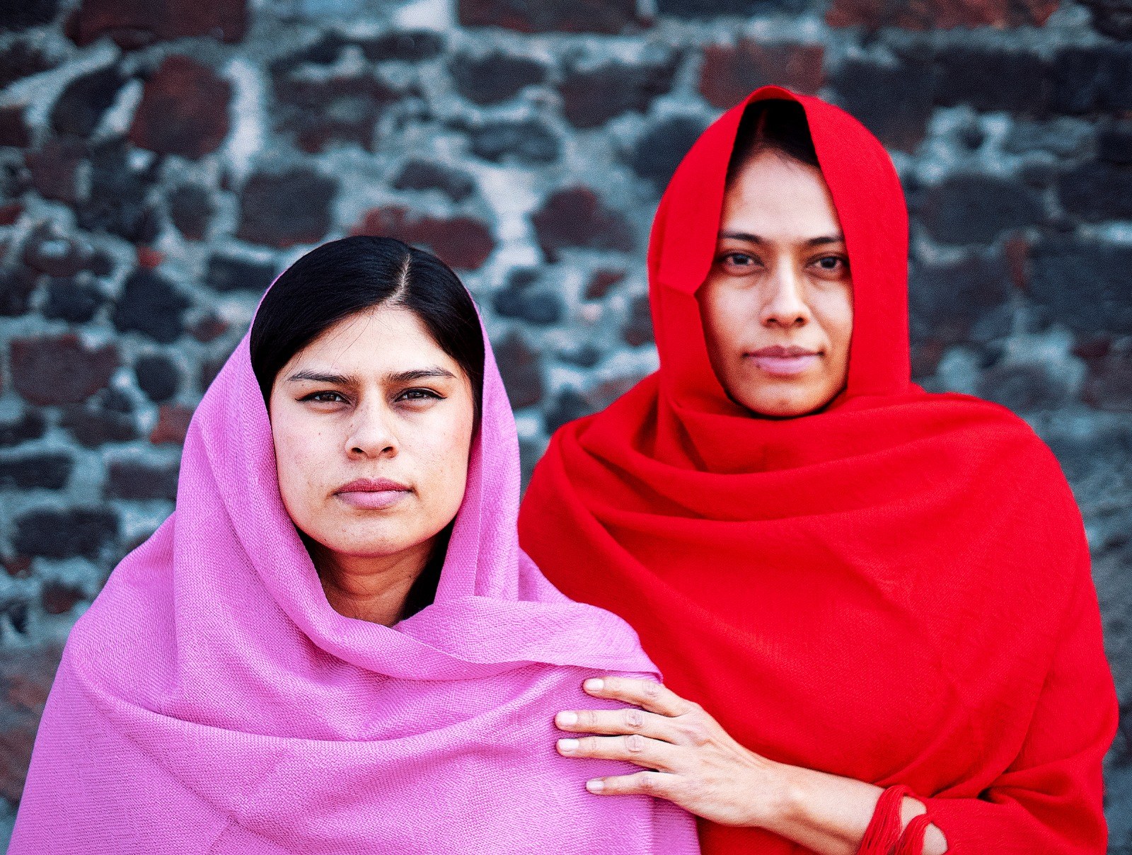 Dos personas retratadas frente a un muro de piedra con telas rosa y roja.